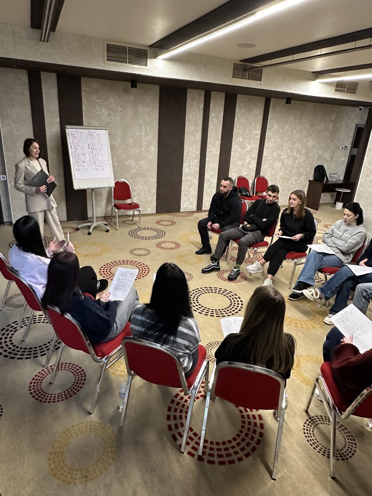 Учасники навчального проєкту сидять у колі в залі з екраном і проектором, конспектують